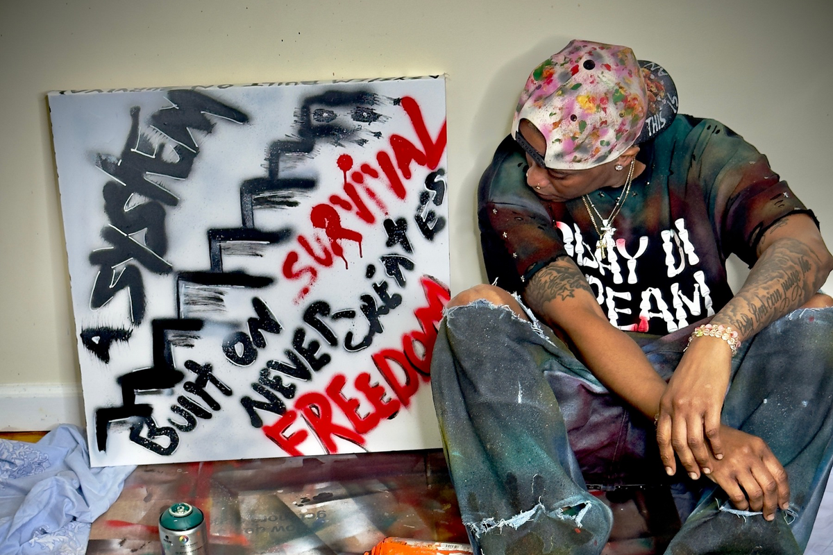 Front view of OBAY DI DREAM's "A System Built on Survival Never Creates Freedom," a black spray-painted staircase with hand-lettered text and red "Survival" and "Freedom," leaning against a wall where the owner sits next to the piece. 