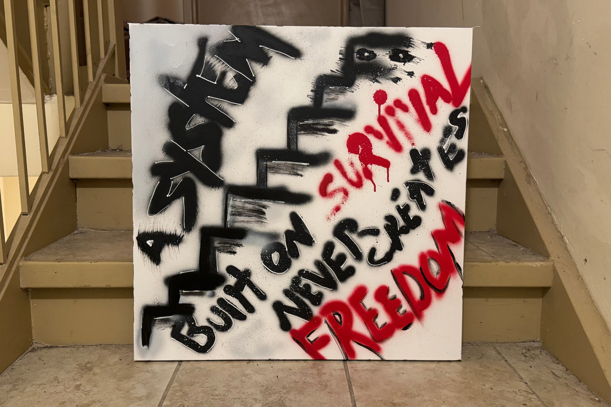 Front view of OBAY DI DREAM's "A System Built on Survival Never Creates Freedom," a black spray-painted staircase with hand-lettered text and red "Survival" and "Freedom," resting at the foot of a stairway.