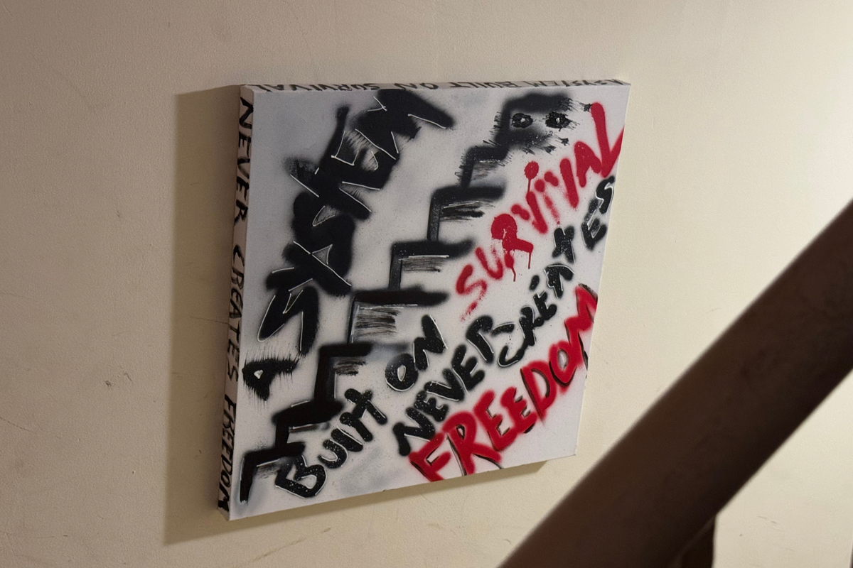 OBAY DI DREAM Unseen Architecture original, "A System Built on Survival Never Creates Freedom," hand-painted in black and red spray paint, hung on a cream wall with the message wrapping around the edges.